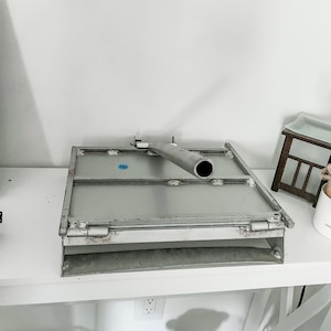 May include: A silver metal industrial-style object with a rectangular base and a hinged lid. A cylindrical pipe extends from the top. The object is on a white surface.