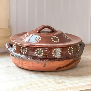 May include: A round, brown clay cooking pot with a matching lid, featuring two side handles and a decorative pattern of small, white-dotted flower motifs encircling both the lid and the upper rim of the vessel, with a smooth, glazed finish.