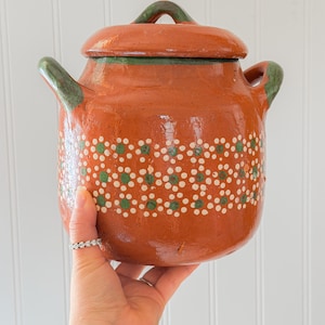 May include: A terracotta clay pot with a matching lid and two side handles. The exterior features a glossy finish with a horizontal band of hand-painted white and green floral patterns. The rim, lid handle, and side handles are accented in green.
