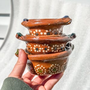 May include: A stack of three small, round, brown clay bowls with two handles each. The exterior of the bowls features a hand-painted pattern of white dots arranged in circular clusters, with occasional green and blue accents on the handles.