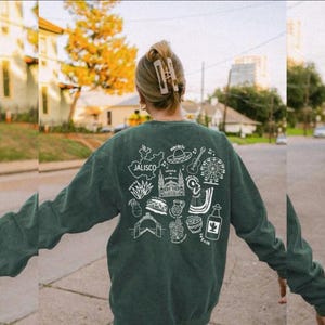 May include: A dark green sweatshirt with a white graphic design on the back featuring the word "Jalisco" and various illustrations of cultural symbols, including a sombrero, guitar, and tequila bottle. The sweatshirt is a solid color and has long sleeves.