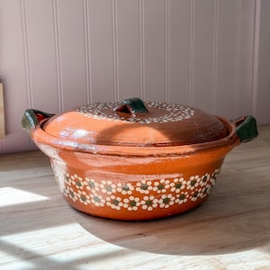 May include: A round terracotta cooking pot with a matching lid, featuring two dark green side handles and a hand-painted pattern of small white and green flowers encircling the base and the top of the lid, with a glossy, smooth finish.