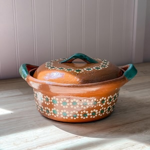 May include: A round, brown clay cooking pot with a matching lid, featuring two side handles and a central lid handle painted in teal. The exterior of the pot and the lid are decorated with a repeating pattern of small white and teal painted flowers.