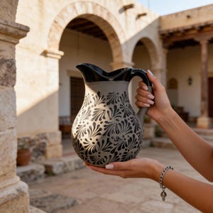 May include: A gray ceramic pitcher with a black rim and handle. The pitcher features a repeating black floral pattern. The pitcher is held in front of a blurred outdoor background.