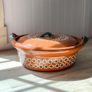 May include: A round terracotta cooking pot with a matching lid, featuring two side handles and a dark green glazed finish on the handles and lid knob. The exterior is decorated with a repeating pattern of small white and green painted flower motifs.