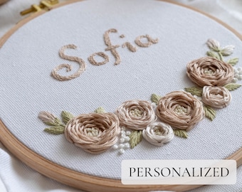 May include: A white embroidery hoop with the name "Sofia" embroidered in brown thread. The hoop is decorated with a floral design of beige and white flowers and green leaves.