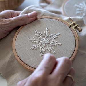 May include: A person is embroidering a white snowflake design on a beige fabric in a wooden embroidery hoop.