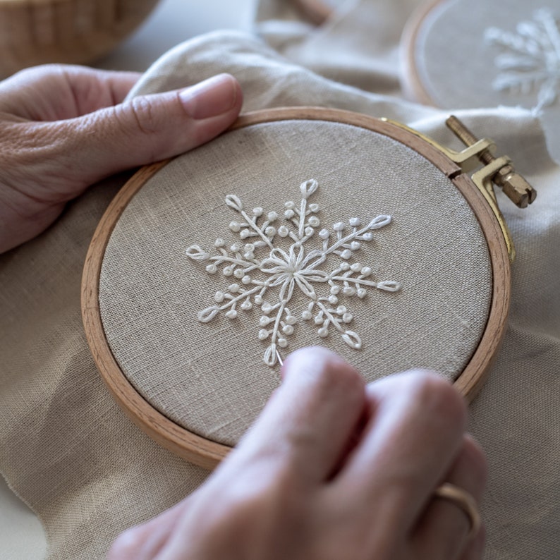 May include: A person is embroidering a white snowflake design on a beige fabric in a wooden embroidery hoop.
