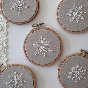 May include: Four embroidery hoops with beige fabric and white snowflake designs. The hoops are different sizes and are arranged in a semi-circle on a white surface.