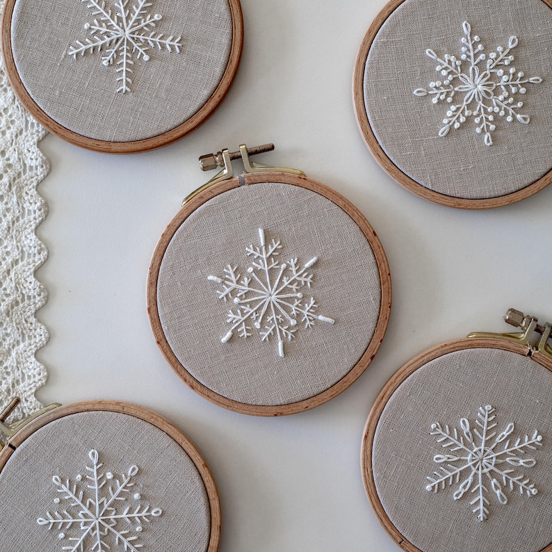 May include: Four embroidery hoops with beige fabric and white snowflake designs. The hoops are different sizes and are arranged in a semi-circle on a white surface.
