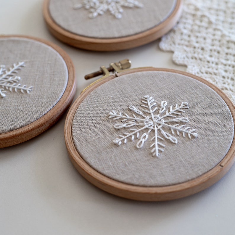 May include: Three embroidery hoops with beige fabric and white embroidered snowflakes. The hoops are made of wood and have a natural finish.