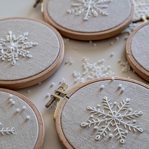 May include: Four embroidery hoops with white snowflake designs on beige fabric. The hoops are arranged in a semi-circle with white beads scattered around them.