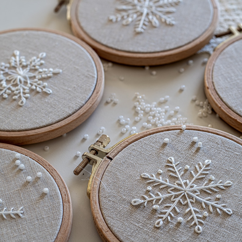 May include: Four embroidery hoops with white snowflake designs on beige fabric. The hoops are arranged in a semi-circle with white beads scattered around them.