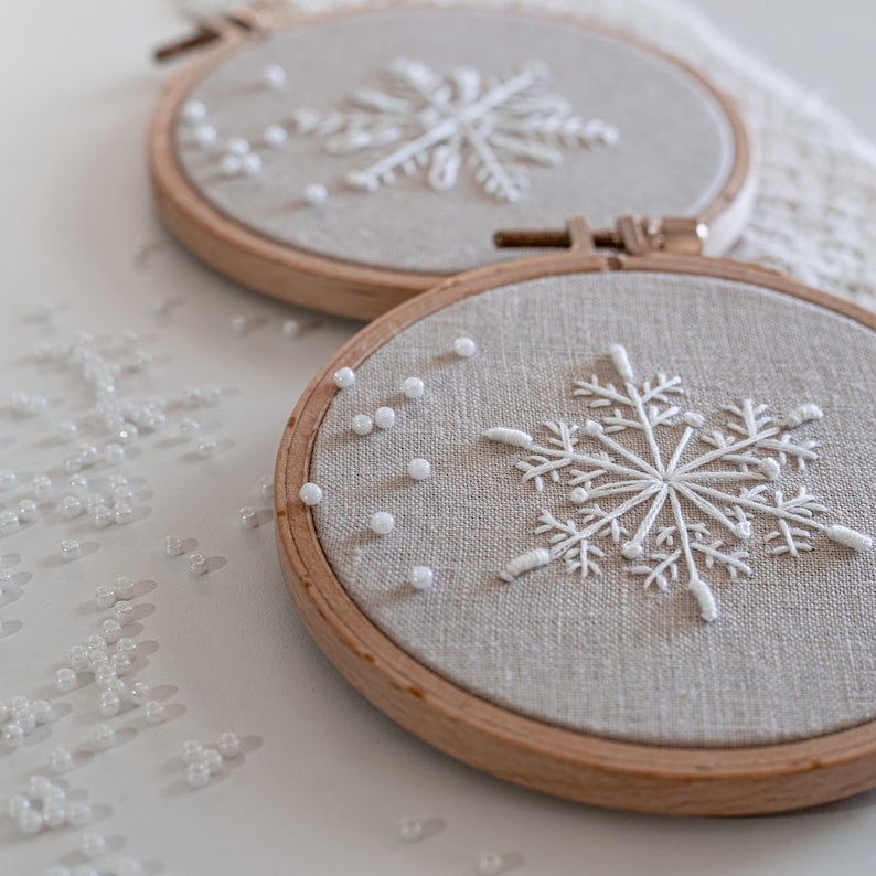 May include: Two embroidery hoops with white snowflake designs on beige linen fabric. The hoops are on a white surface with white beads scattered around them.