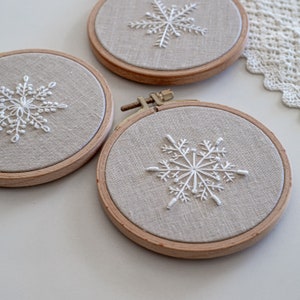 May include: Three embroidery hoops with white snowflake designs on beige linen fabric. The hoops are made of natural wood and have a simple, elegant design.