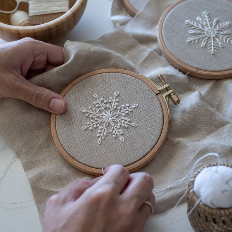 May include: A person is embroidering a white snowflake design on a beige fabric stretched in a wooden embroidery hoop. The hoop is on a beige fabric surface.