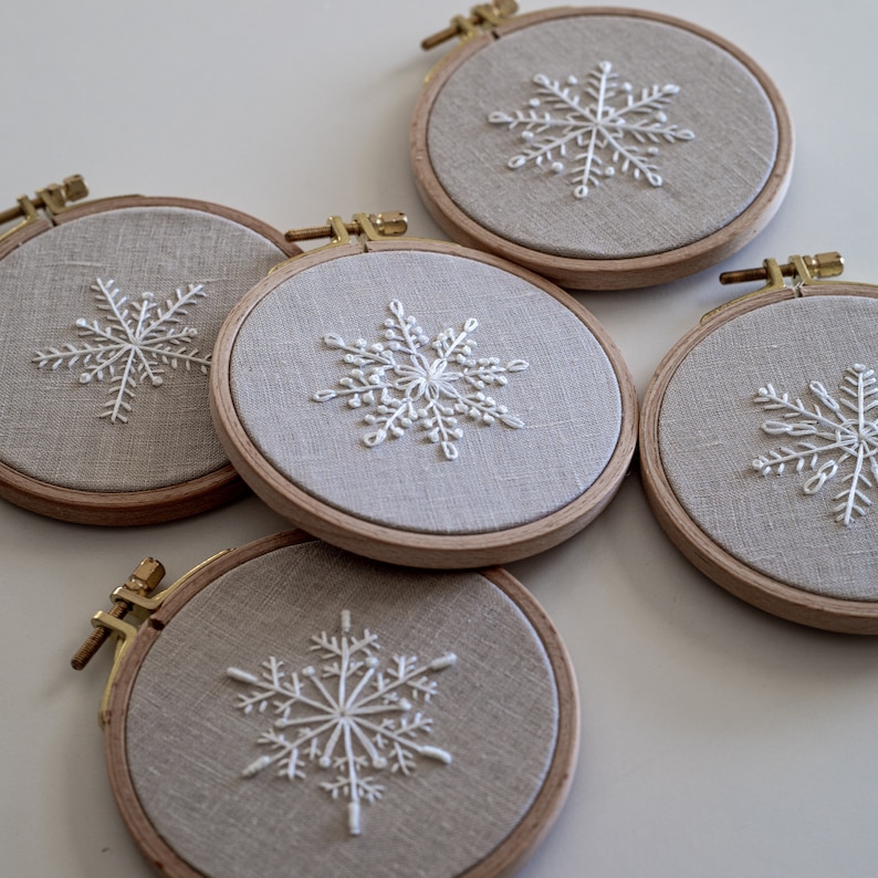 May include: Four embroidery hoops with white snowflake designs on beige linen fabric. The hoops are made of wood and have a gold metal ring.