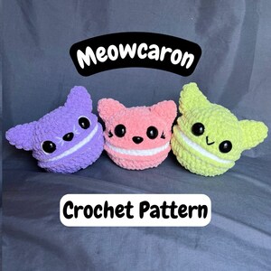 May include: Three crocheted cat-shaped macarons in purple, pink, and green. The macarons have white frosting and black eyes. The text "Meowcaron Crochet Pattern" is below the macarons.