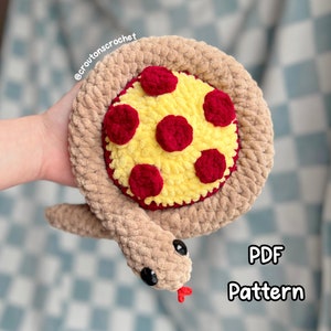 May include: A crocheted pizza snake toy with a brown body and a red and yellow pizza topping. The snake is curled up in a circle with its head facing the camera. The text "PDF Pattern" is visible in the image.
