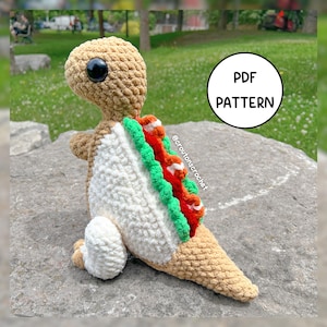 Puede incluir: Un dinosaurio de ganchillo en beige y blanco, con una espalda en forma de taco. El dinosaurio tiene un ojo negro grande y un diseño de concha de taco verde, rojo y blanco. La imagen incluye un círculo blanco con el texto "PDF PATTERN".