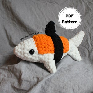 Könnte beinhalten: Ein weißer gehäkelter Hai mit orangefarbenen und schwarzen Details, der einem Sushi-Rollen ähnelt. Der Hai hat ein schwarzes Auge und eine kleine schwarze Flosse oben. Der Text "PDF Pattern" ist in einem Kreis auf dem Bild geschrieben.