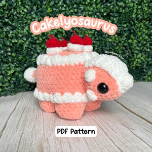 May include: A crocheted pink and white cake dinosaur with red cherries on top. The dinosaur is sitting on a wooden surface. The text "Cakelyosaurus" is written in pink letters above the dinosaur. The text "PDF Pattern" is written below the dinosaur. The text "@crou tonscrochet" is written in small letters on the right side of the dinosaur.