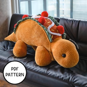 May include: A crocheted stuffed toy shaped like a dinosaur with a taco shell on its back. The toy is brown and orange with red and green accents. The toy is sitting on a black leather couch. The text "PDF PATTERN" is visible in the lower left corner of the image.
