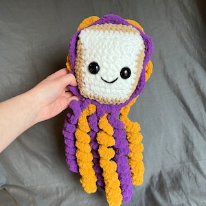 May include: A crocheted octopus plush toy with a white square head, brown and purple yarn for the body, and yellow and purple yarn for the tentacles.