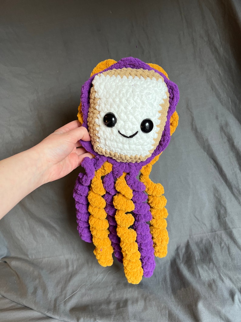 May include: A crocheted octopus plush toy with a white square head, brown and purple yarn for the body, and yellow and purple yarn for the tentacles.