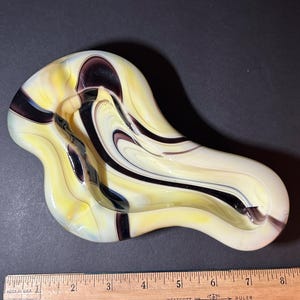 May include: An irregular, kidney-shaped glass dish featuring a marbled swirl pattern of cream, pale yellow, and deep purple colors. A wooden ruler is positioned along the bottom edge, showing the object measures approximately 9.5 inches in length.