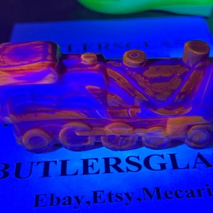 May include: A glass train ornament in shades of orange and purple, set against a blue backdrop. The text "BUTLERSGLASS" and "Ebay, Etsy, Mecari" are visible. The train features a detailed design.