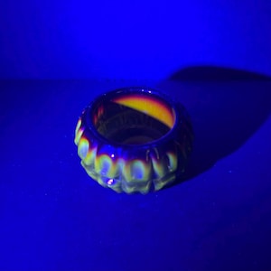 May include: A small, circular glass bowl featuring a repeating vertical ribbed pattern along its exterior. The glass exhibits a distinct iridescent glow with yellow and green highlights against a dark, deep purple background under ultraviolet light.