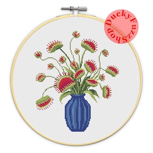 May include: Cross-stitch of a Venus flytrap bouquet in a blue vase. The red and green flytraps are in a blue vase, set against a white background. The cross-stitch is displayed in a wooden embroidery hoop.