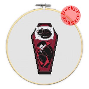 May include: A cross-stitch pattern in a round wooden embroidery hoop featuring a pixel art design of a vampire and a black cat resting inside a dark red coffin. The text DuckyFuzzShop is printed in red in the upper right corner of the white fabric.