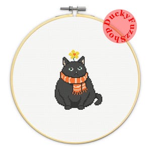 May include: A cross-stitch embroidery in a round wooden hoop featuring a plump black cat wearing an orange and white striped scarf with a small yellow flower on its head. The design is centered on white fabric with a DuckyFuzzShop logo in the corner.
