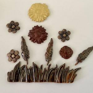 May include: A collection of ceramic decorative elements in various shapes and colors. The set includes flowers, feathers, and grass in shades of brown, tan, and yellow. The pieces are arranged on a white surface.