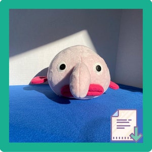 May include: A plush toy blob fish with a light pink body, large black eyes, and bright pink lips and fins. The toy is sitting on a blue surface, with a white and gray background. The toy is a whimsical and unique item.