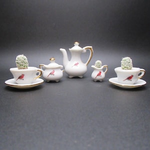 May include: A white porcelain tea set with gold trim and a red bird design. The set includes a teapot, sugar bowl, creamer, and two teacups with saucers, each holding a small cactus plant.