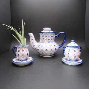 May include: A miniature ceramic tea set with a teapot, creamer, and cup and saucer. The set features a pink and blue floral pattern with blue trim. A small plant sits in the cup. The set is displayed on a dark surface.