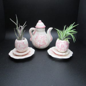 May include: A white ceramic teapot and two matching planters, each holding a small plant. The teapot and planters are decorated with a pink floral pattern and pink trim. The set is displayed on a black background.