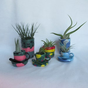 May include: A collection of colorful ceramic planters, including teacup-shaped and cylindrical designs, each holding small green plants. The planters feature various patterns and colors, such as polka dots, stripes, and swirls, creating a whimsical aesthetic.
