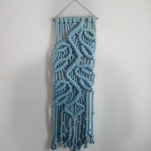 May include: A light blue macrame wall hanging with a leaf-like design. The handmade textile features intricate knotting patterns and hanging fringe. The wall decor is suspended from a metal rod and cord, adding a touch of bohemian style to any space.