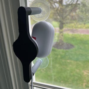 May include: A black and gray window mount for a security camera. The mount uses suction cups to attach to the window. A white security camera is attached to the mount. A white cable is connected to the camera.