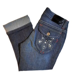May include: Blue denim cropped jeans with a decorative pocket featuring a rhinestone design and red stitching. The back pocket has a black leather patch with the text "Rock & Republic" in white.