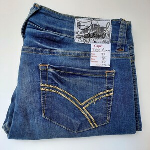 May include: Blue denim capri jeans with a back pocket featuring a gold embroidered design. The jeans have a tag with the text "Capri Code Denim" and size details: Size 29, Waist 30", Rise 8", Inseam 16".