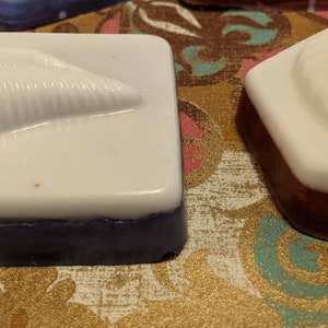 May include: Two handmade soap bars with a seashell design. One bar is white with a seashell imprint and a dark blue base. The other bar is white with a seashell imprint and a brown base.