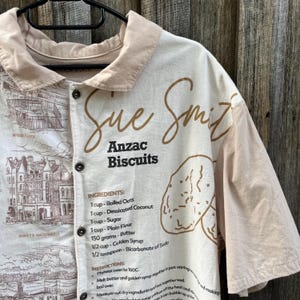 May include: A short-sleeved button-down shirt in cream and beige featuring a brown graphic print of an Anzac Biscuits recipe, including ingredients and instructions, alongside a detailed sketch of a building labelled Barry's Amusements.