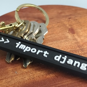 May include: Black keyring with white text that reads ">>> import django". The keyring is attached to a gold key ring with several keys.
