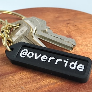 Puede incluir: Llavero negro con un texto blanco '@override' sobre un fondo negro. El llavero est&aacute; unido a un juego de llaves con un llavero dorado.
