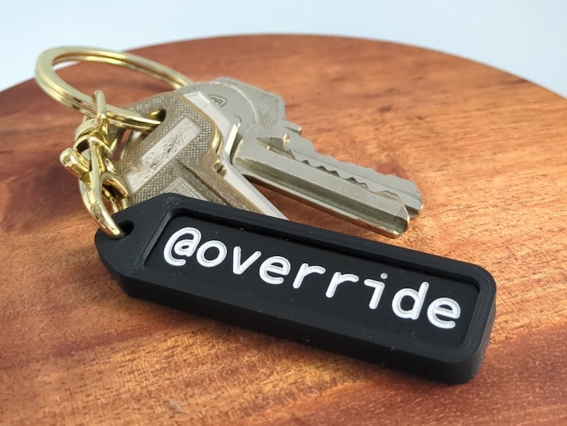 Puede incluir: Llavero negro con un texto blanco '@override' sobre un fondo negro. El llavero est&aacute; unido a un juego de llaves con un llavero dorado.